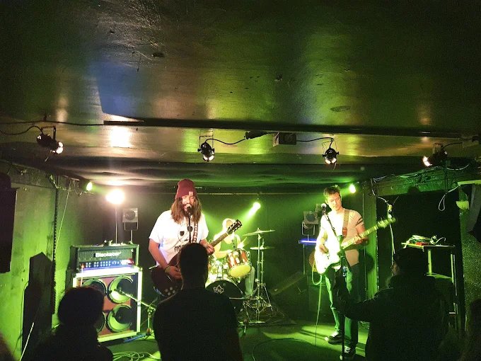 Un groupe de rock joue sous des lumières vertes dans la petite salle de Leda Atomica, ampli et batterie au fond